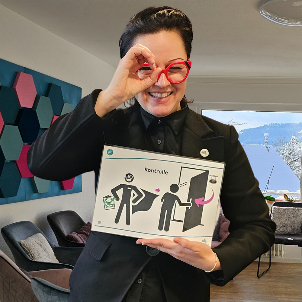 Diyana steht in einem Raum, schwarz gekleidet mit roter Brille, lächelt in die Kamera und macht mit einer Hand eine Kreisform mit dem Daumen und Zeigefinger, durch die sie mit einem Auge durchblickt. In der anderen Hand hält sie ein Schild mit zwei Figuren, die eine Figur öffnet eine Tür und blickt hinein